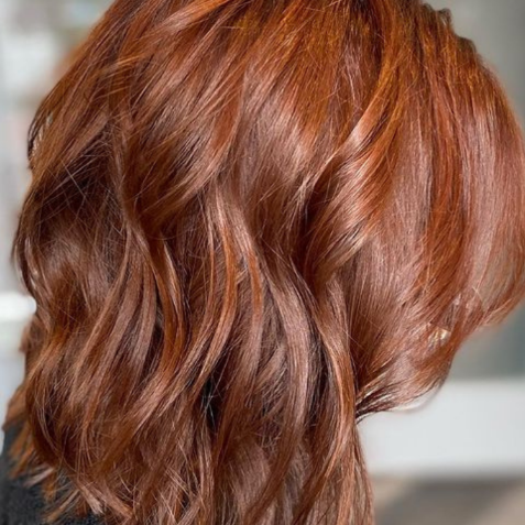 Close-up of a person with wavy, shoulder-length red hair, viewed from the side, with a blurred background suggesting a hair salon setting.