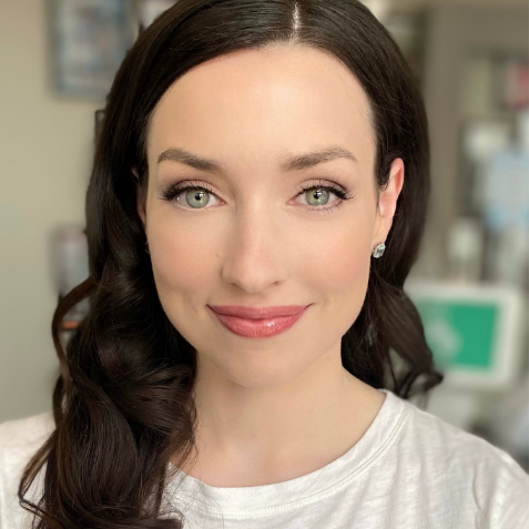 A woman with long dark hair styled in loose curls, wearing subtle makeup and a white shirt, smiles at the camera in a well-lit indoor setting.